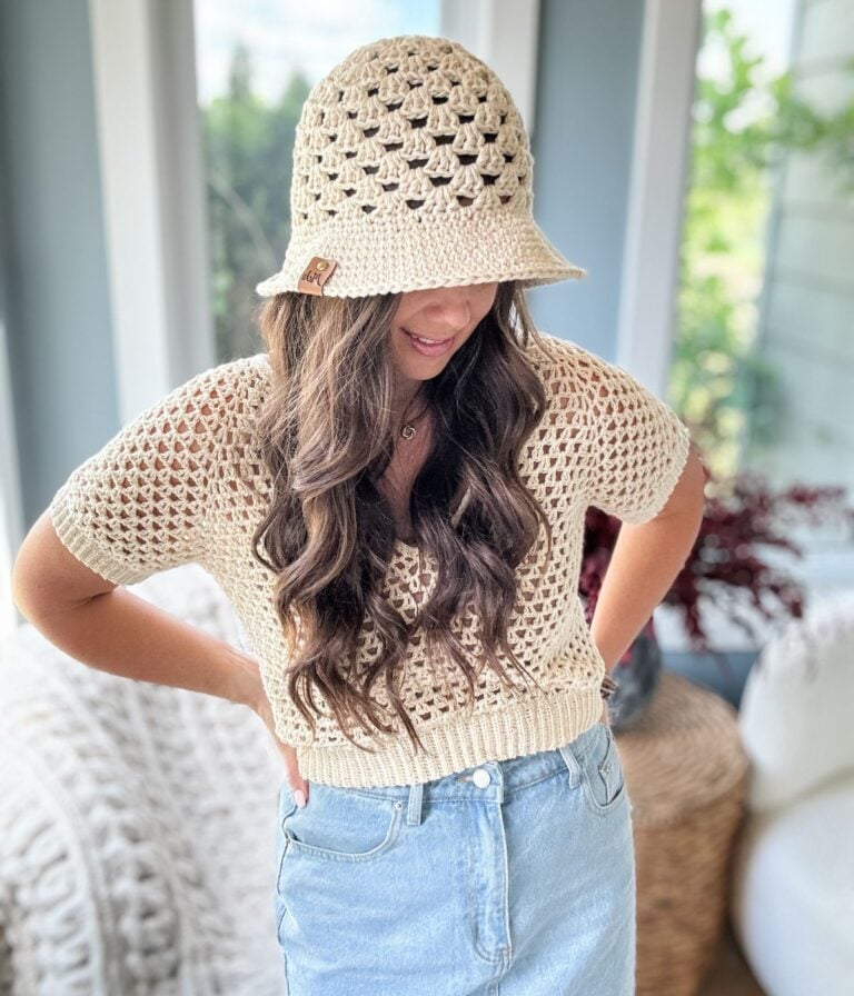 A woman with long hair wears a beige crochet top and matching hat, paired with a light blue denim skirt. She stands indoors near a window, her hands resting on her hips.