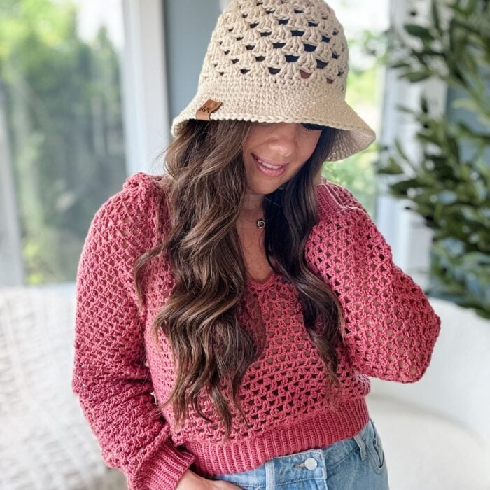 A person with long hair wearing a crochet hat and pink crochet top stands indoors, looking down.