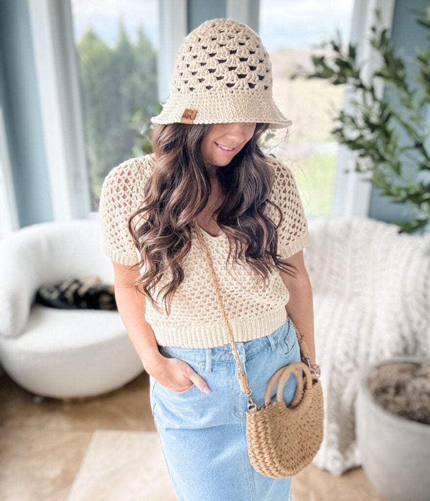 A person stands indoors wearing a beige crochet top, paired with a matching bucket hat and a woven crossbody bag, and complemented by a light blue denim skirt. In the background, there’s a white chair next to a green plant.