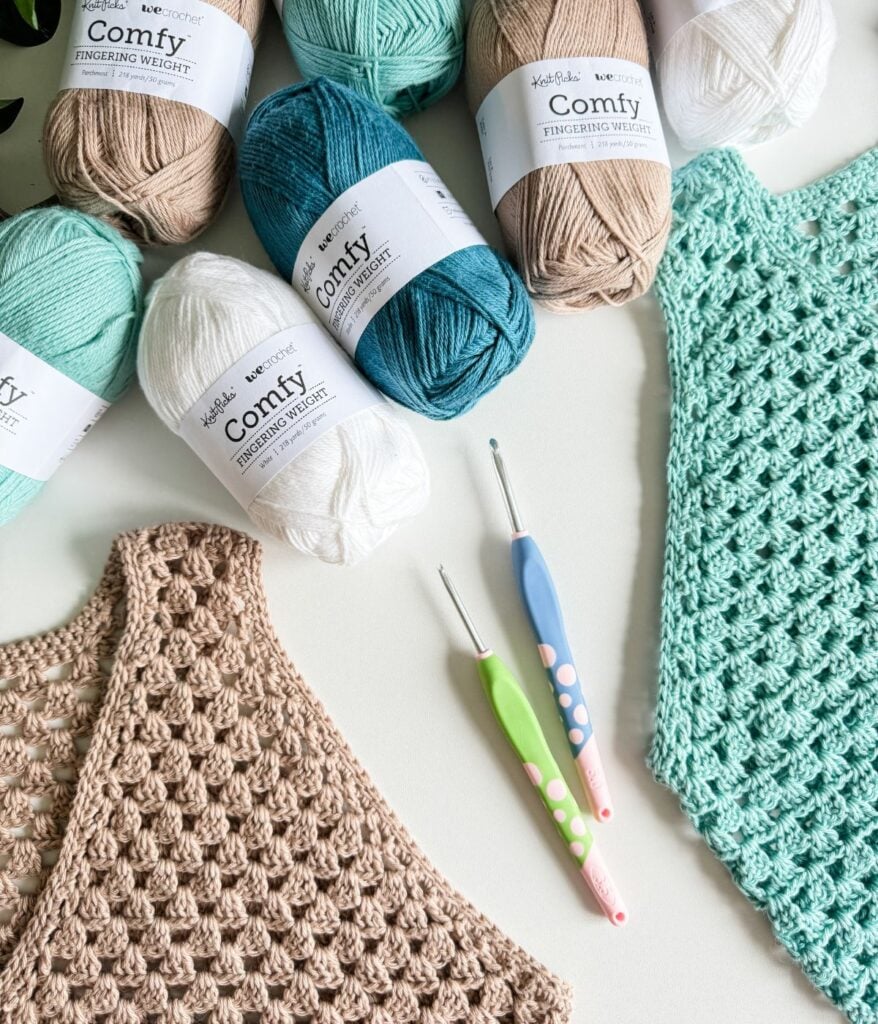 Assorted skeins of yarn in white, teal, brown, and off-white arranged beside two crochet hooks and two crocheted tank tops—one in beige and one in teal, both featuring a charming granny stitch. Find your next favorite free crochet pattern to create your own stylish crochet tank top.