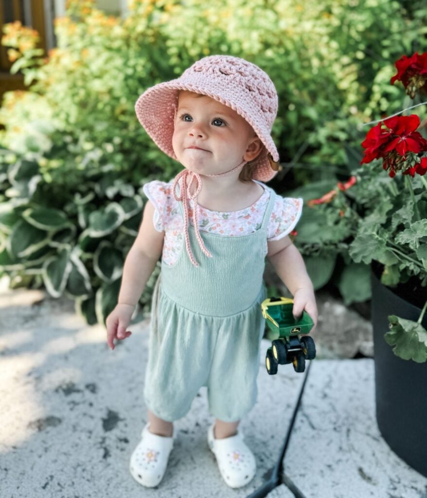 A toddler wearing a pink hat, green overalls, and a cute crochet top holds a toy tractor, standing outside on a sidewalk beside green plants and red flowers.