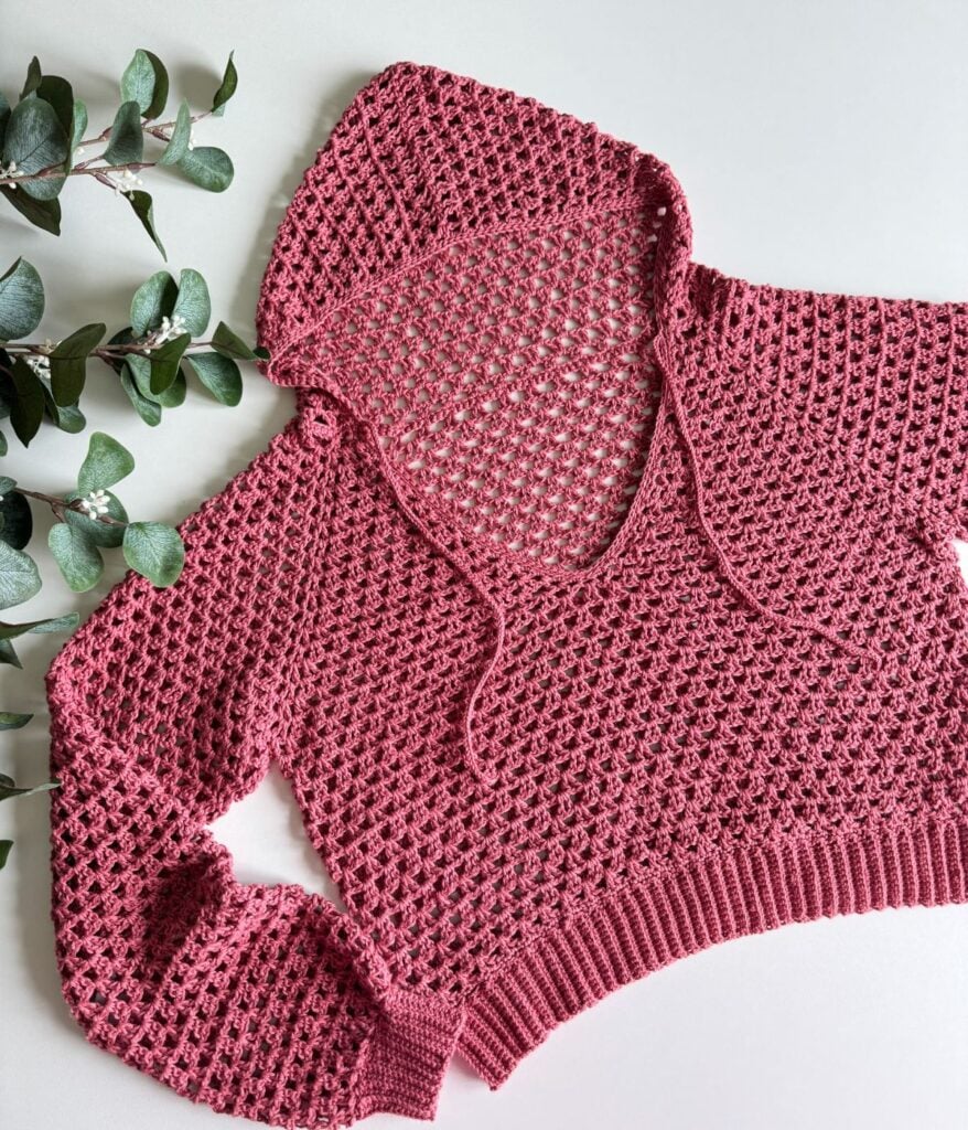 A pink, knit sweater with a hood and drawstrings is laid flat on a white surface next to green leaves, resembling the cozy charm of a handmade crochet top.