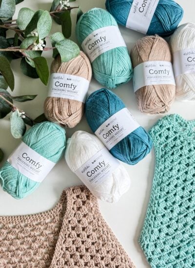 Various skeins of KnitPicks Comfy yarn in shades of blue, green, white, and beige are arranged alongside two crochet projects using the Easy Granny Stitch and some eucalyptus leaves.