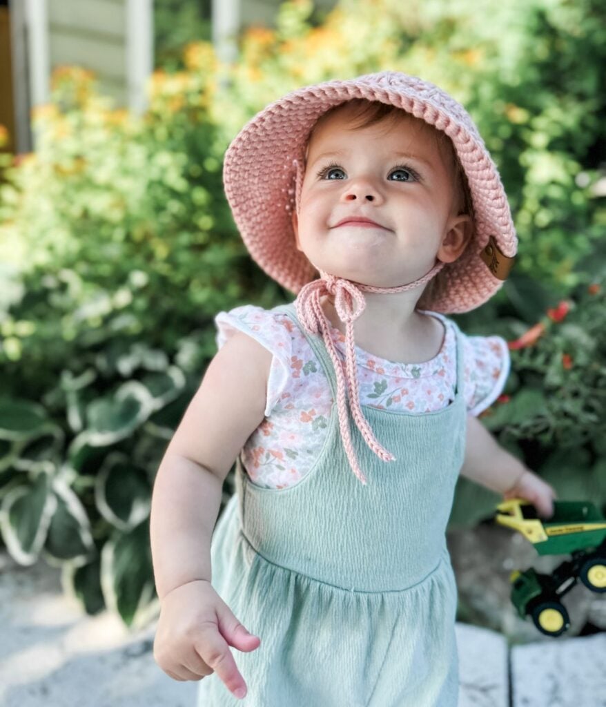 A toddler wearing a pink hat and a green dress with a crochet top looks up while holding a toy tractor. There are green plants and flowers in the background.