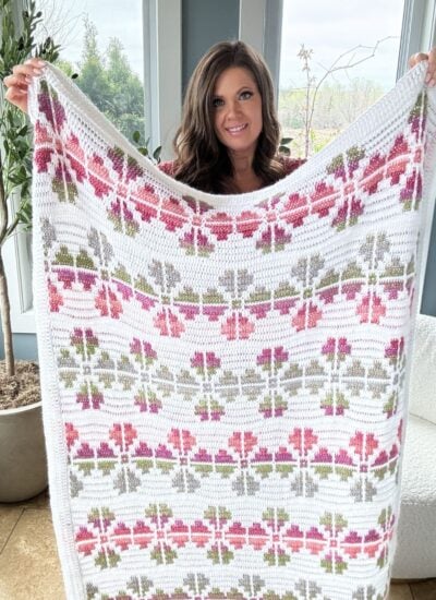 A woman with long brown hair holds up a Flower Blanket featuring a pattern of red, pink, and green flowers against a white background. She stands indoors with a plant and a large window nearby, showcasing her beautiful Cosmos Mosaic Crochet work.