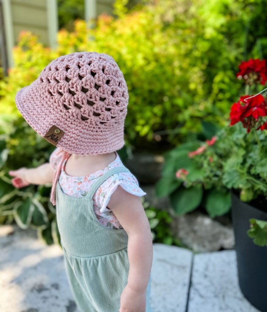 A toddler wearing a pink crocheted hat and green overalls stands in a garden with red flowers and lush greenery, looking like a tiny gardener in their adorable crochet top.