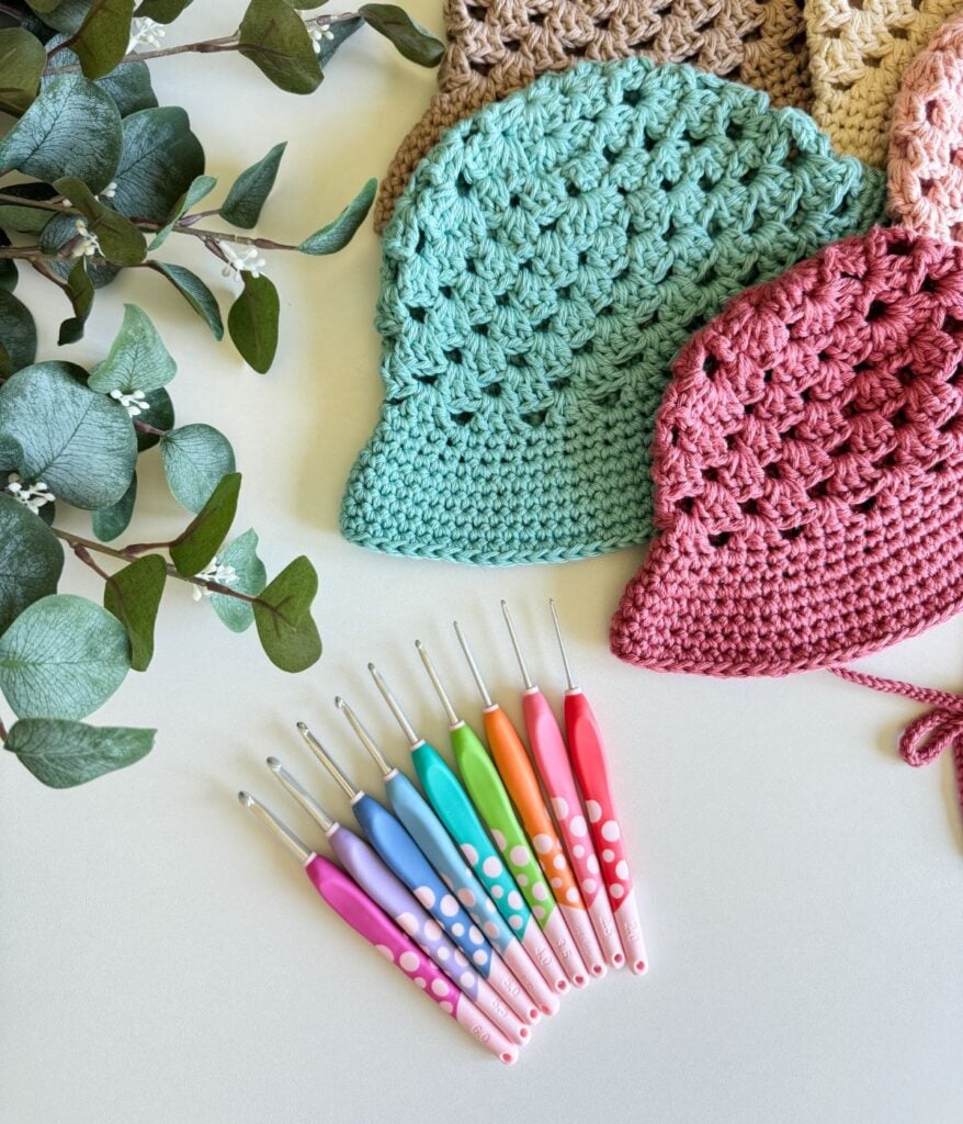 A set of colorful crochet hooks is laid out on a table next to green leaves, several knitted hats in various colors, and a vibrant crochet top.