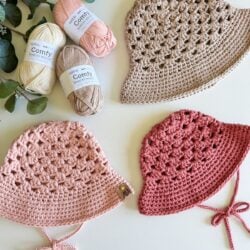 Three crochet hats in beige, pink, and red are displayed alongside a stylish crochet top on a surface adorned with four skeins of multi-colored yarn and green leafy branches.