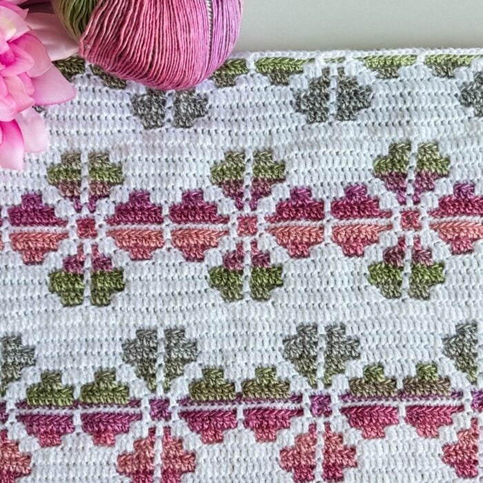A colorful crochet pattern with pink, green, and purple geometric shapes, resembling a Cosmos Mosaic. A skein of pink and green yarn and a delicate crochet flower are partially visible at the top left corner.