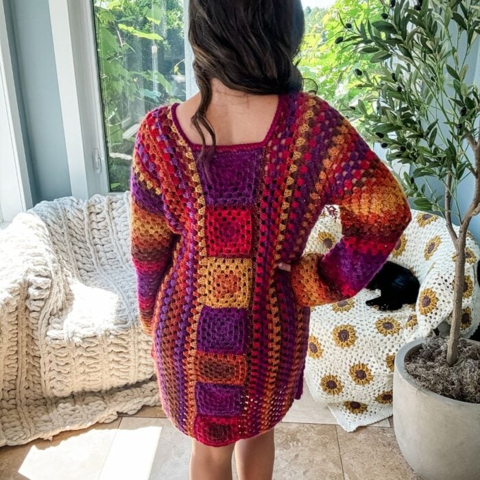 A person with long dark hair is standing indoors with their back to the camera, wearing a colorful crocheted sweater-dress made from a Granny Stitch free crochet pattern. Nearby are a knitted blanket and a large potted plant, giving the scene a charming retro vibe.