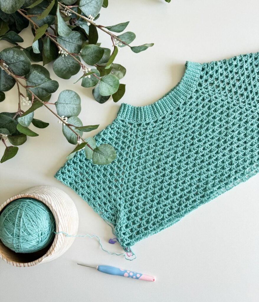 A teal crocheted piece, a ball of matching yarn in a bowl, a crochet hook, and eucalyptus branches are arranged on a light surface, hinting at the beginnings of a summer cardigan.