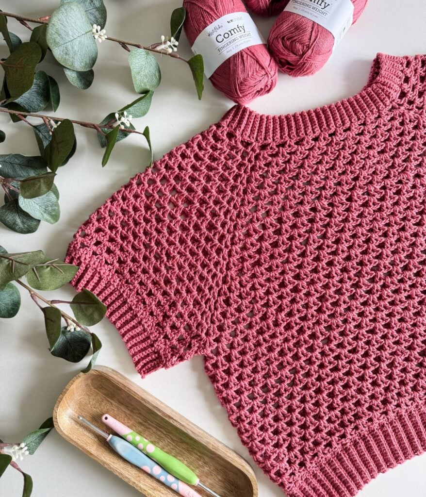A knitted pink summer cardigan is displayed on a table with three skeins of pink yarn, two crochet hooks in a wooden tray, and eucalyptus branches.