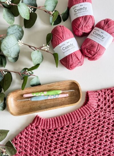 Crocheted pink summer cardigan, made with three skeins of "Comfy Fingering Weight" pink yarn, crochet hooks in a wooden tray, and eucalyptus branches arranged on a flat surface.
