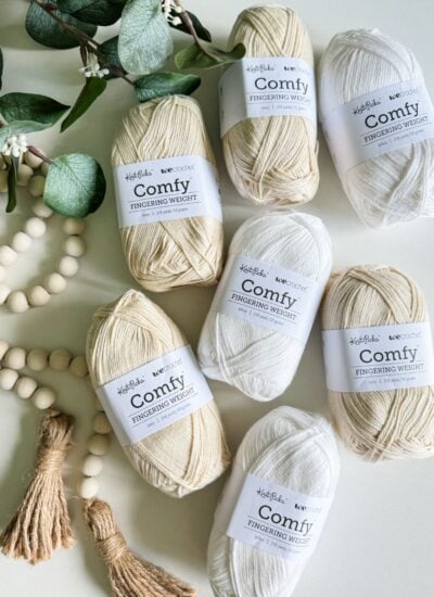 Several skeins of "Comfy Fingering Weight" yarn in shades of beige and white are arranged on a surface with a wooden bead garland and green foliage, perfect for crafting a cozy crochet cardigan.