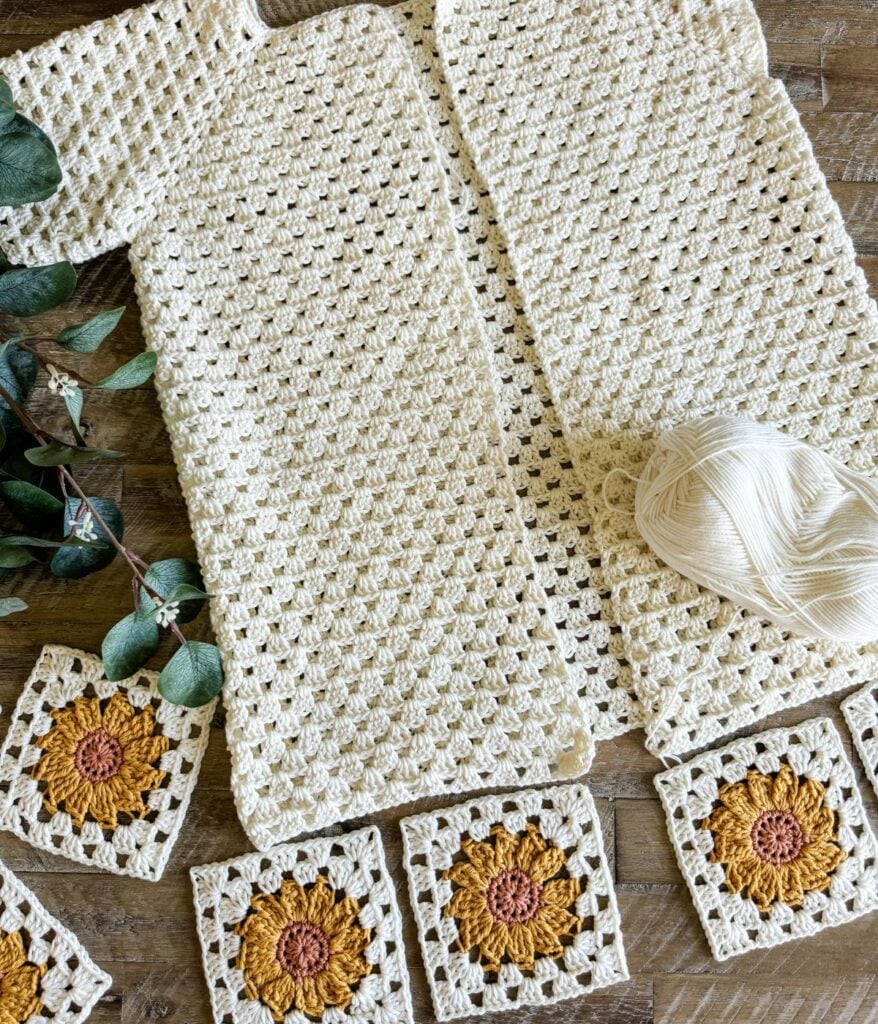 A crocheted white blanket in progress lies on a wooden surface with a ball of white yarn and a crochet hook. Sunflower crochet squares, reminiscent of a sunflower cardigan, are beautifully placed around the blanket edges.