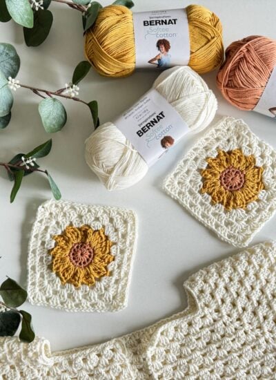 Two crochet sunflower squares and a partially completed Sunflower Cardigan are displayed with three balls of Bernat Softee Cotton yarn in yellow, white, and peach colors, alongside some green leafy branches.