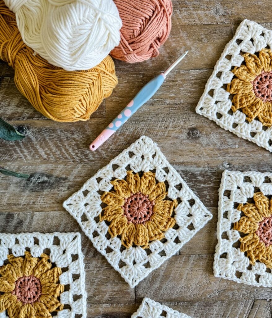 Crocheted sunflower squares are laid out on a wooden surface with balls of yarn and a crochet hook, perfect for crafting your own sunflower cardigan.