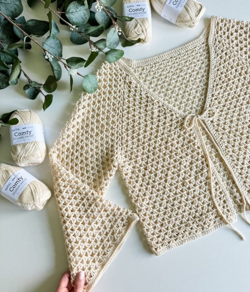 A hand holding the sleeve of a crocheted beige cardigan placed on a table alongside several balls of beige "Comfy" yarn and green eucalyptus branches.
