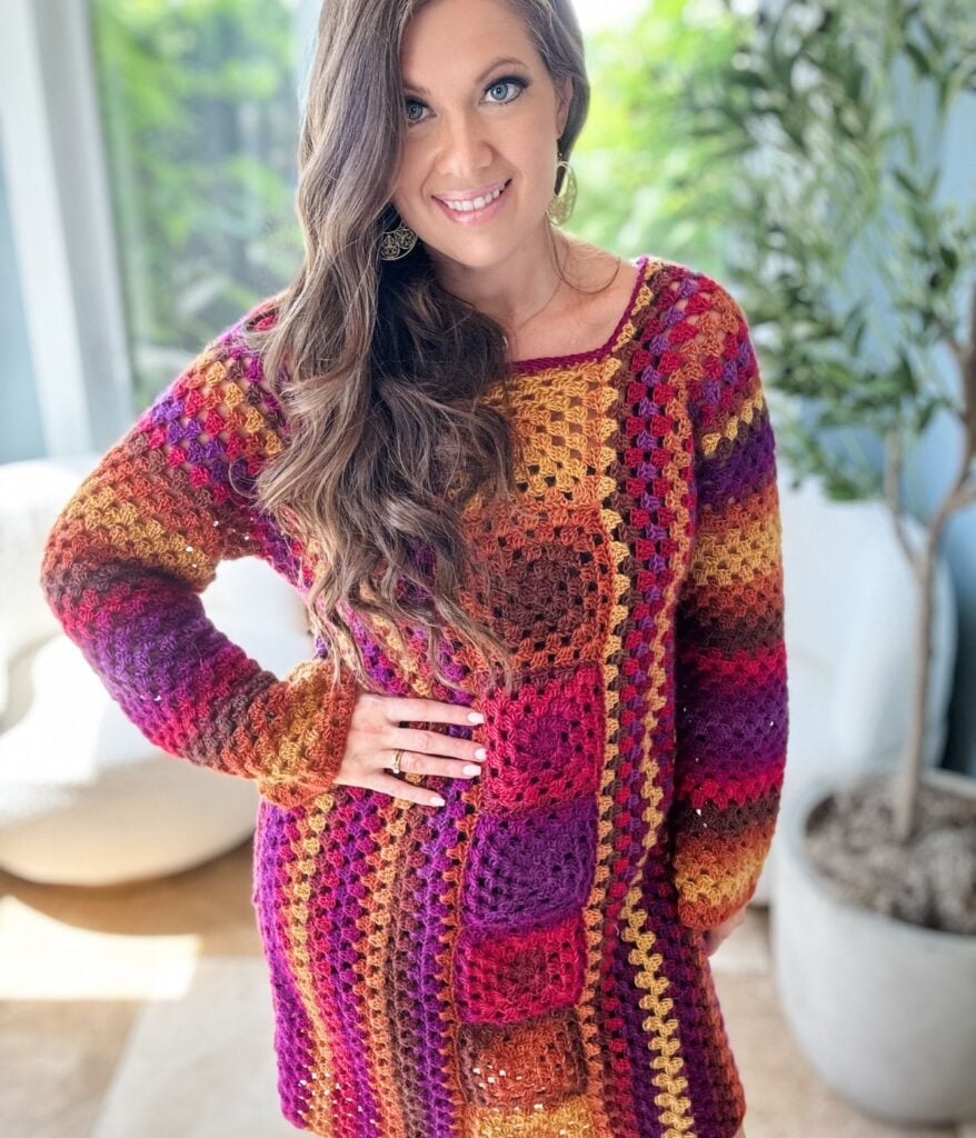 A woman with long brown hair stands indoors wearing a colorful crochet sweater. She is smiling and has her hand on her hip. A plant is visible in the background, reminiscent of a Retro Granny Stitch Dress.