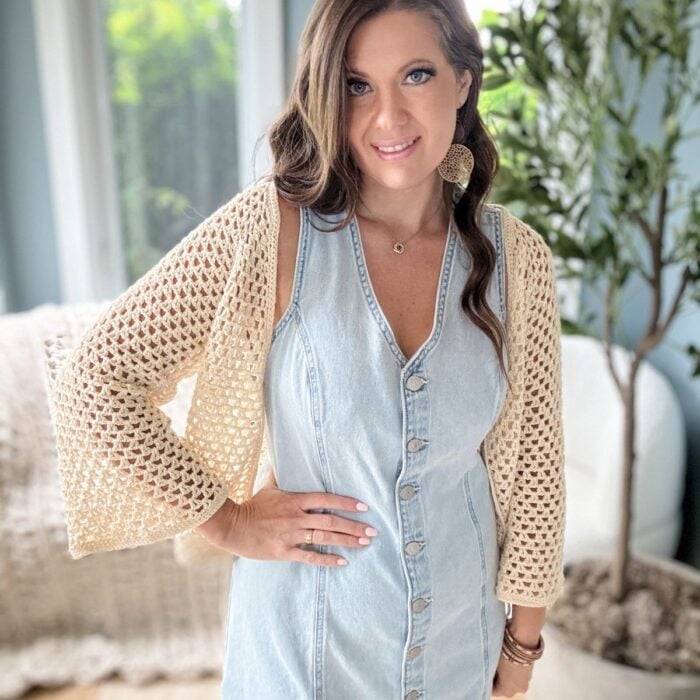 A woman in a denim button-front dress and a crochet cardigan stands indoors next to a plant, with her right hand on her hip. She has long brown hair and is wearing large earrings.