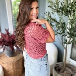 A person with long brown hair stands indoors, looking over their shoulder and smiling. They wear a pink crochet summer cardigan and denim jeans. There are plants and wicker baskets in the background.