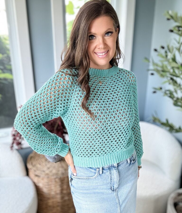 A woman standing indoors, wearing a light blue crochet summer cardigan over a light-colored top and denim skirt, with her hand on her hip, looking at the camera and smiling.