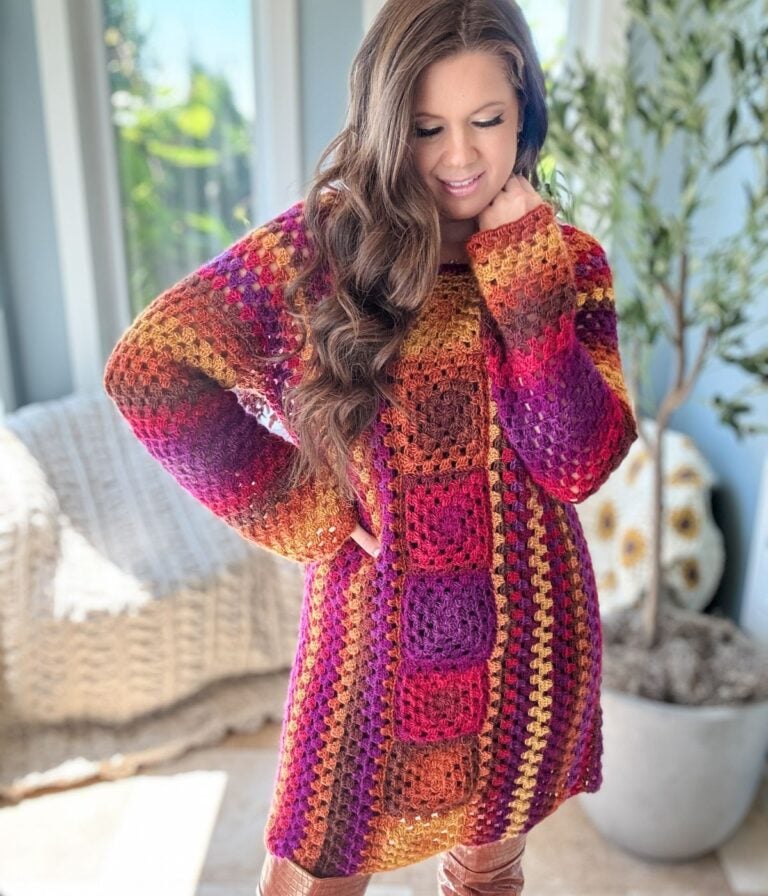 A person with long, wavy hair is wearing a colorful, striped Retro Granny Stitch Dress. They are standing indoors near a window with greenery outside and a plant in the background.
