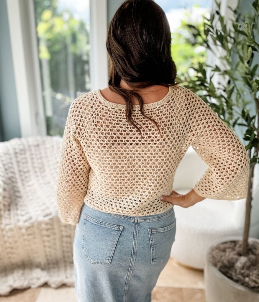 A woman with long brown hair, seen from behind, stands indoors wearing a beige crochet cardigan and blue denim jeans. There is a green plant to her right and a large window in the background.