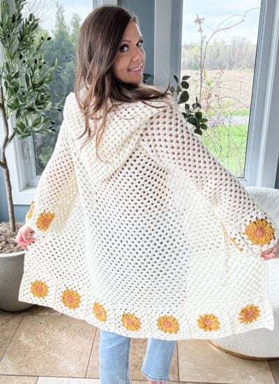 A woman smiles, showcasing the back of a beautiful sunflower cardigan adorned with intricate yellow flower designs near the hem. She stands indoors by a window, her warmth and joy reflected in both her expression and the exquisite craftsmanship of her attire.