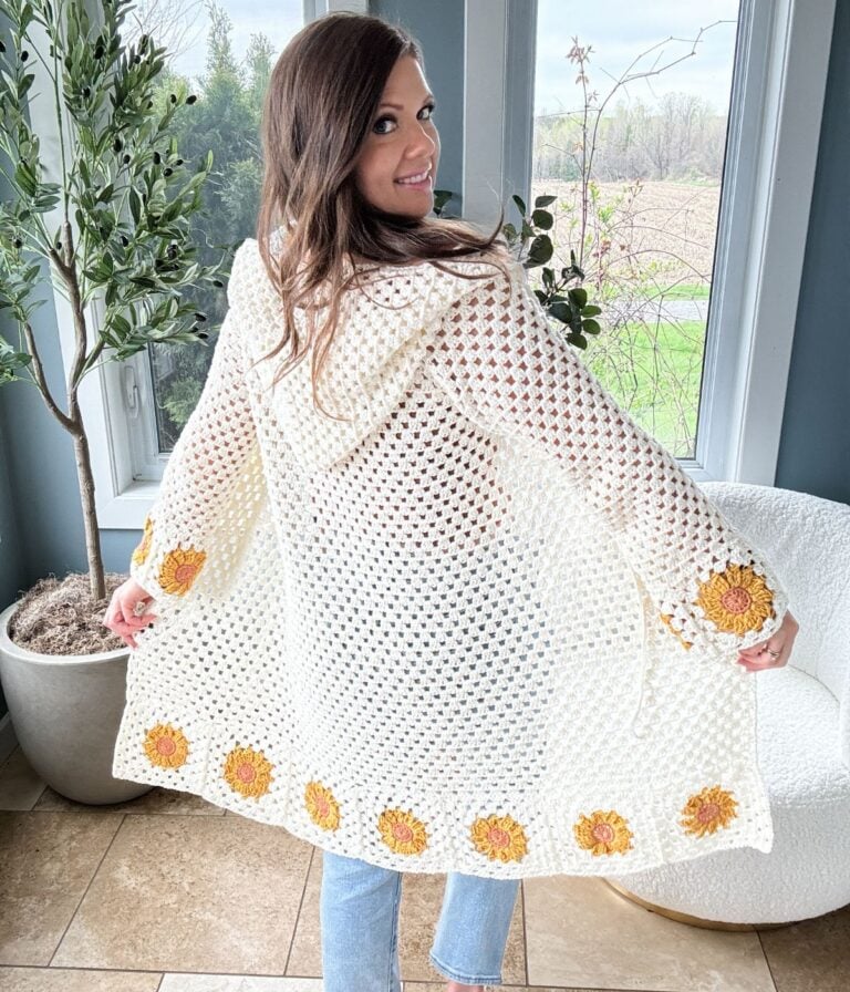 A woman smiles, showcasing the back of a beautiful sunflower cardigan adorned with intricate yellow flower designs near the hem. She stands indoors by a window, her warmth and joy reflected in both her expression and the exquisite craftsmanship of her attire.