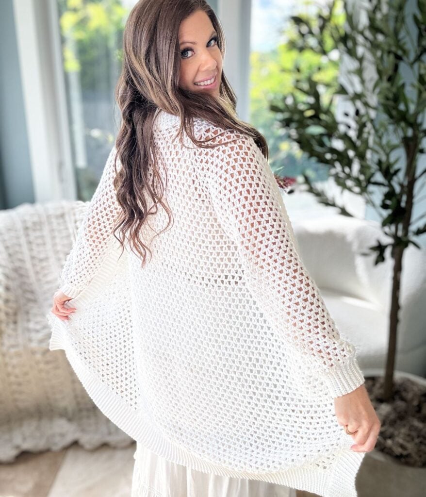 A person with long hair smiles and turns slightly, showcasing a white, open-knit crochet cardigan in a well-lit room with a plant and furniture in the background.