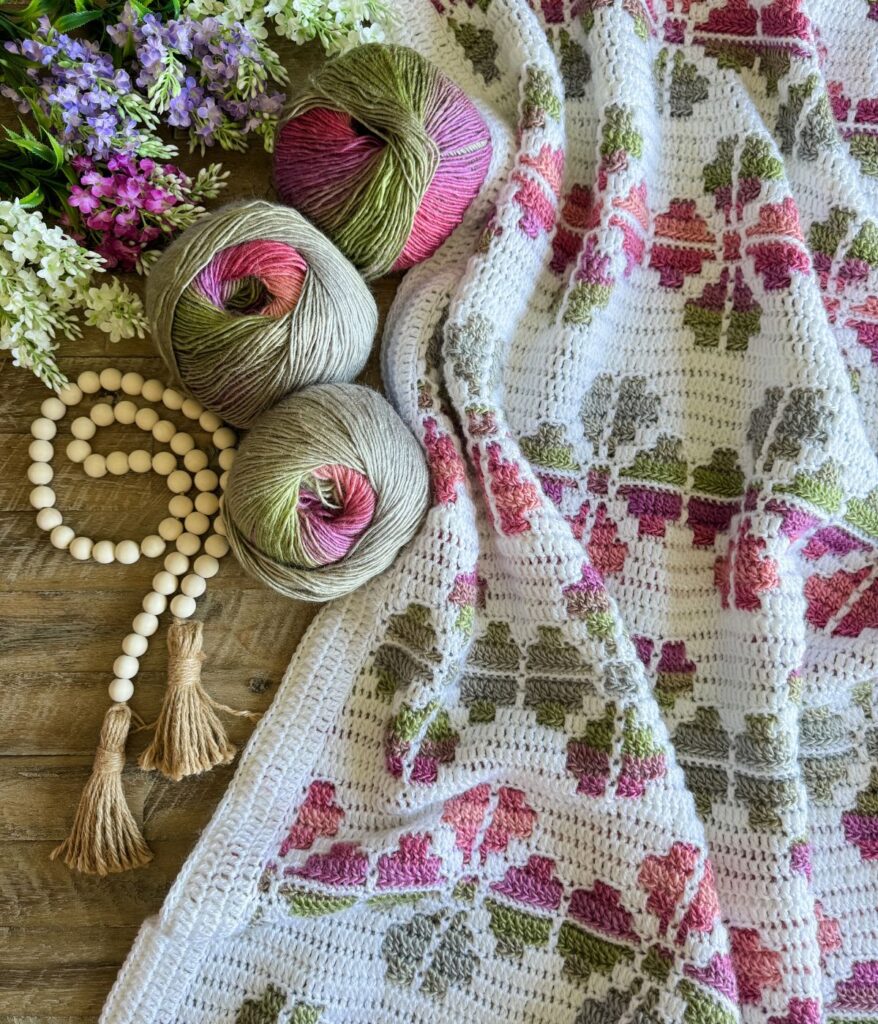 A Mosaic Crochet blanket with a floral pattern in pink, green, and gray yarn, three yarn balls, wooden beads with tassels, and artificial purple and white flowers laid out on a wooden surface.
