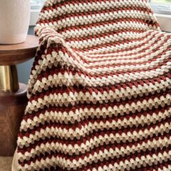 A red, brown, and beige striped Granny Stitch Blanket is draped over a chair near a wooden side table with a white vase.