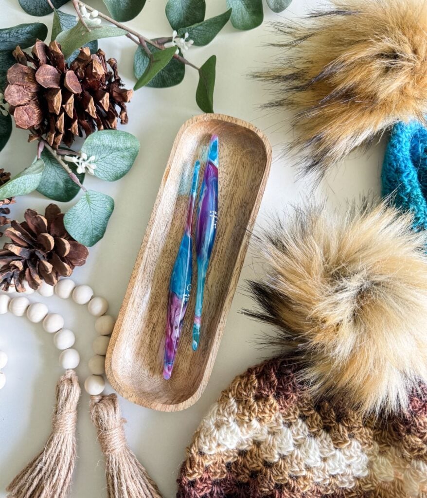 Two colorful crochet hooks placed in a wooden tray, surrounded by pine cones, leaves, a beaded tassel, and Granny Stitch Hats with furry pom-poms.