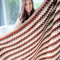 A woman smiles while holding up a large Granny Stitch Blanket with a striped pattern in shades of brown, cream, and red.