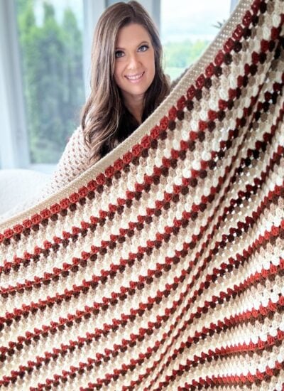 A woman smiles while holding up a large Granny Stitch Blanket with a striped pattern in shades of brown, cream, and red.
