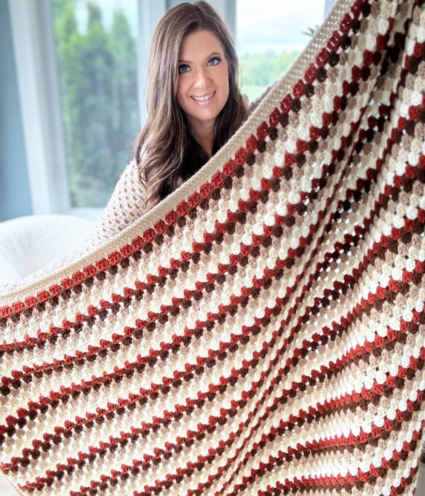 A woman smiles while holding up a large Granny Stitch Blanket with a striped pattern in shades of brown, cream, and red.