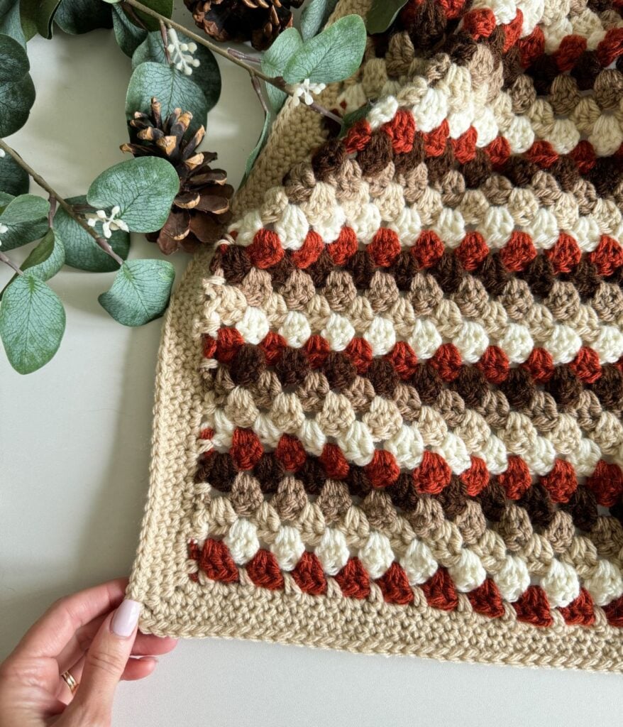 A hand holds the corner of a Granny Stitch Blanket with a striped pattern in brown, beige, and orange. Pinecones and eucalyptus leaves are placed beside it.