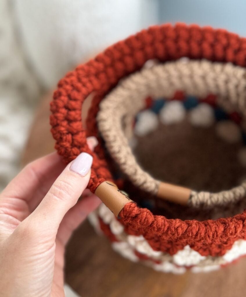A hand holds the edge of a red and beige Granny Stitch Basket with leather detail, placed inside another crocheted basket.