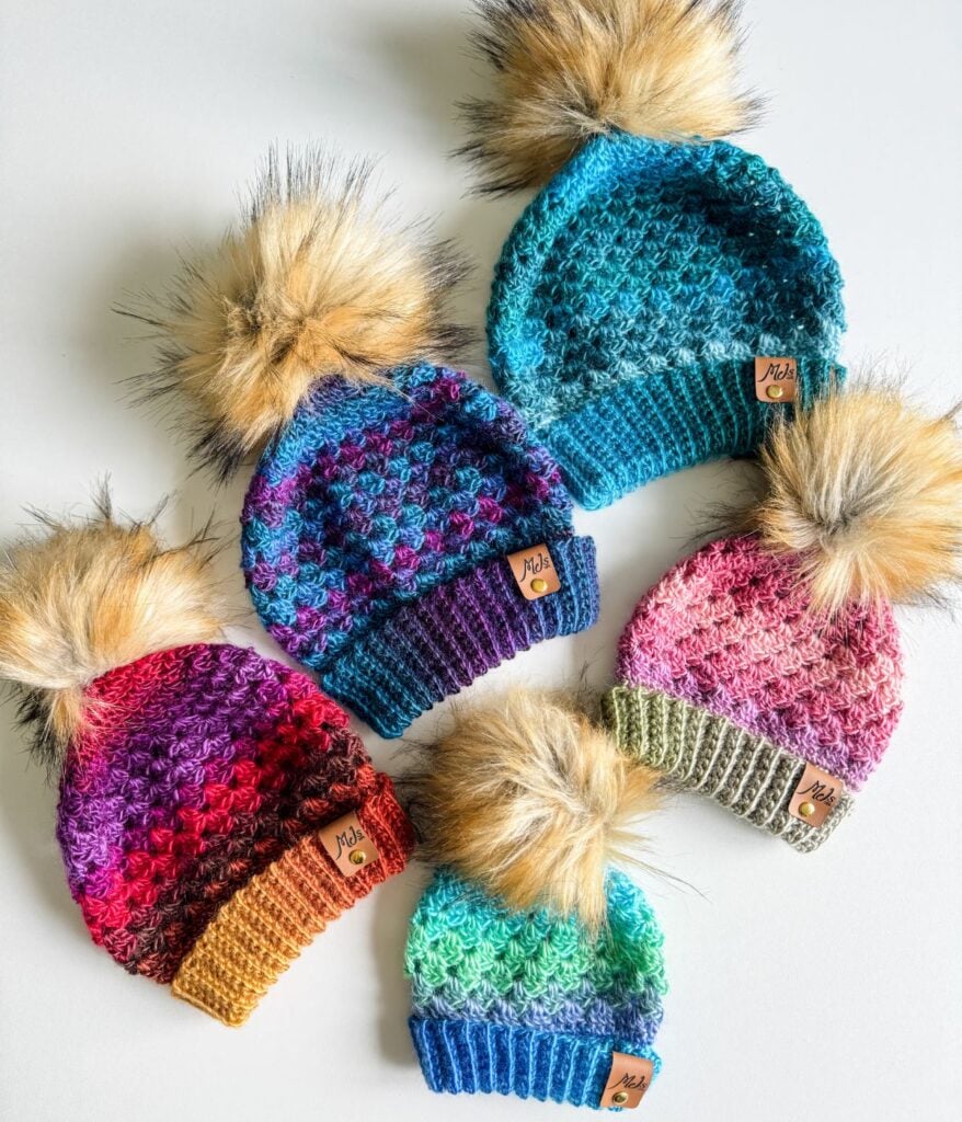Five colorful Granny Stitch hats with faux fur pom-poms are arranged in a flat lay. Each beanie features a unique color pattern and a small leather tag on the brim.