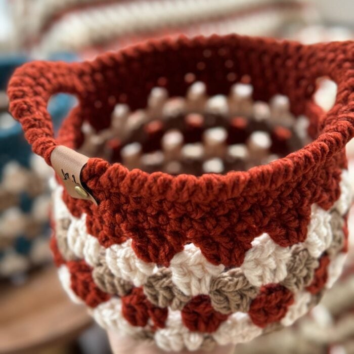 A hand holds a beautifully handcrafted Granny Stitch Basket, featuring a red rim and an alternating red, white, and tan pattern.
