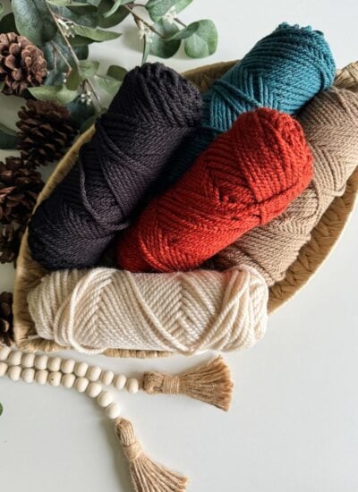 Granny Stitch Basket containing five skeins of yarn in various colors, surrounded by pinecones, eucalyptus leaves, and a tassel bead garland.