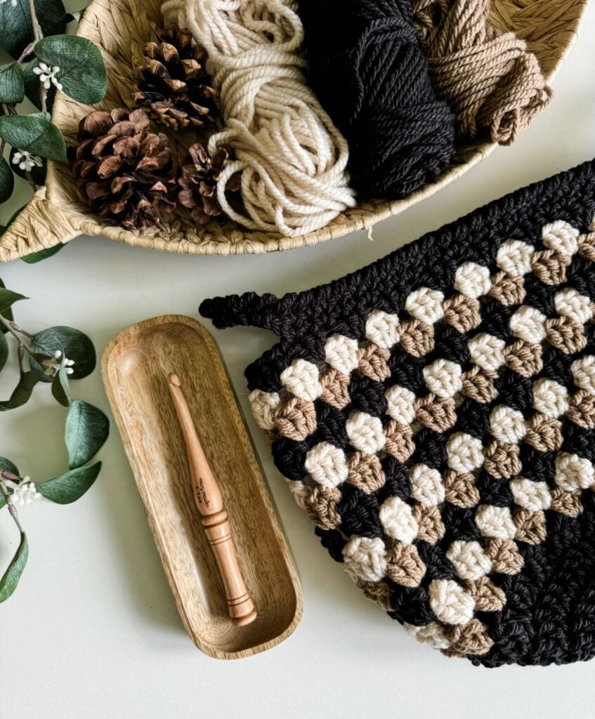 Granny Stitch Basket project in progress with yarn balls, a wooden crochet hook, pinecones, and greenery on a table.