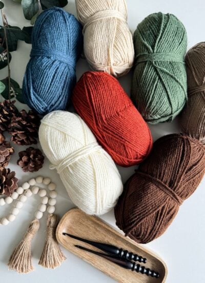 A collection of yarn skeins in various colors are arranged on a flat surface alongside pinecones, beads, wooden tassels, and crochet hooks, perfect for creating a charming granny stitch pumpkin or another delightful crochet pattern.