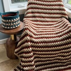 A cozy armchair is covered with a Granny Stitch Blanket in red, brown, and cream stripes, next to a small wooden table with a matching crocheted basket on it.