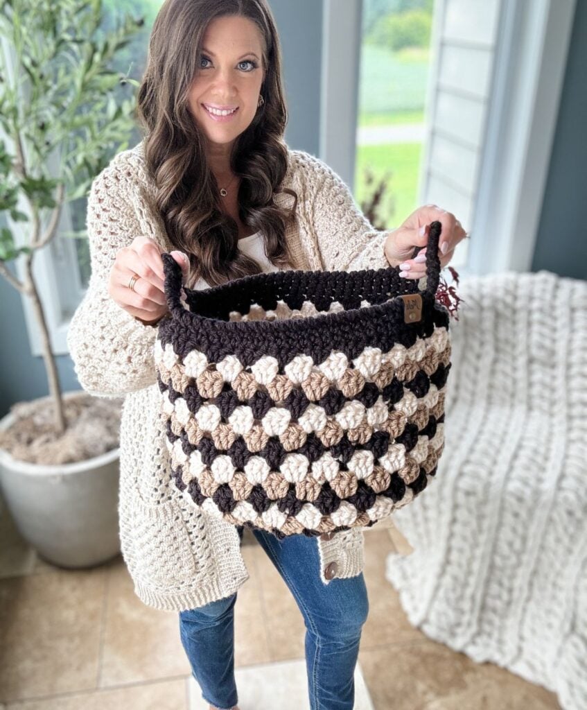A woman smiling and holding a black and beige Granny Stitch Basket in a room with a potted plant and a knitted blanket draped over a chair.