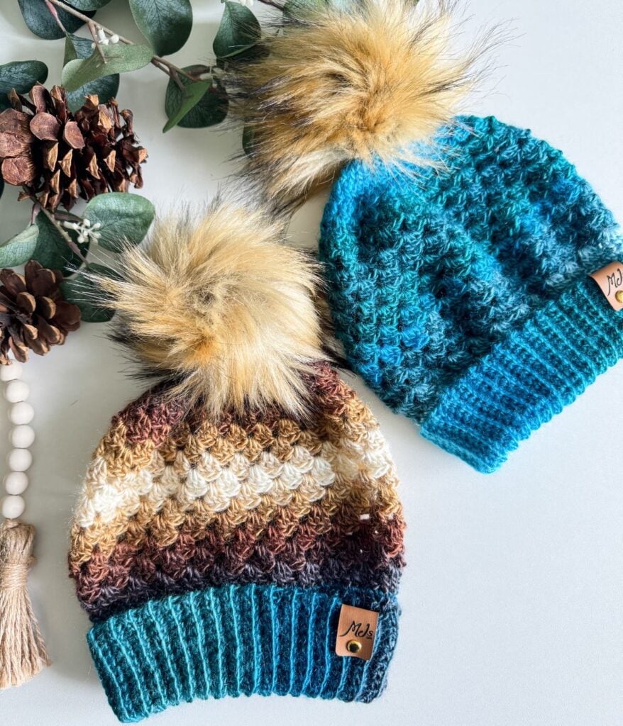 Two knit beanies with large fur pom-poms rest on a surface—one in multicolored stripes resembling a Granny Stitch Hat and the other in solid teal. Pinecones and eucalyptus leaves are placed beside them.