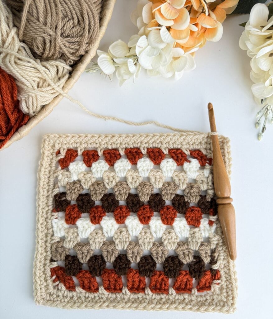 A crocheted granny stitch blanket in shades of white, brown, beige, and orange is surrounded by balls of yarn, a wooden crochet hook, and some white and orange flowers.