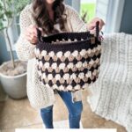 A woman holding a Granny Stitch Basket in beige and black colors, standing indoors next to a window and a potted plant. Text below reads "FALL FOR GRANNY BASKETS Crochet pattern + Video".
