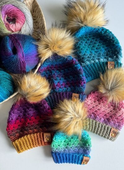 A collection of colorful Granny Stitch hats with faux fur pom-poms, arranged on a table, next to several skeins of yarn in a woven basket.